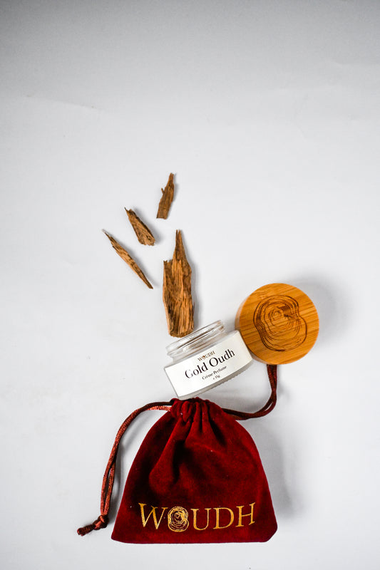 Agarwood chips and Gold Oudh solid perfume in a reusable glass container labeled Gold Oudhl' on a red velvet drawstring bag with 'WOUDH' branding.