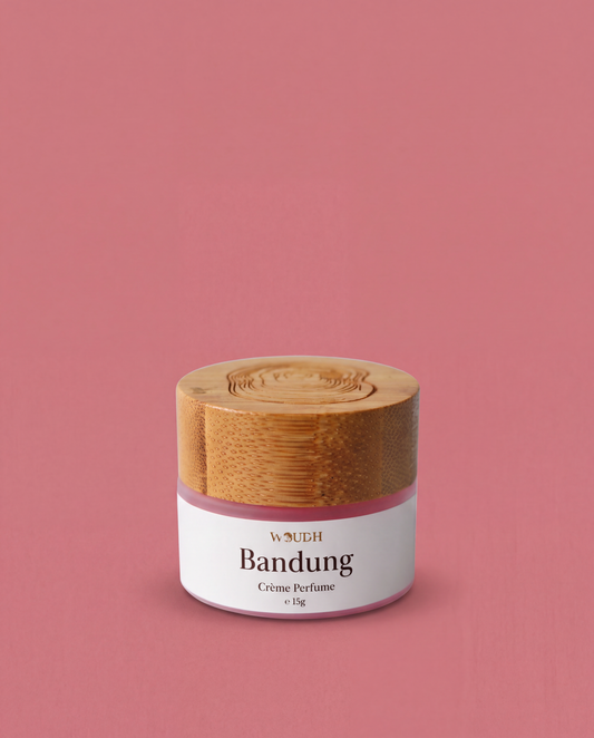 A frosted glass jar labeled 'WOUDH Bandung Crème Perfumé' with a white label and a light wood-grain lid, centered on a solid dusty pink background.