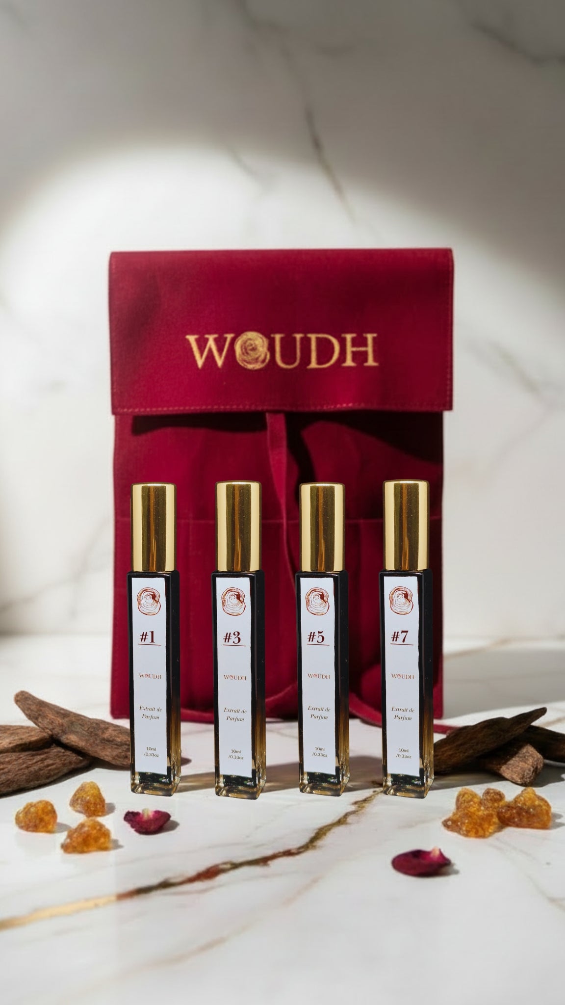 Four WOUDH perfume spray bottles with gold caps lined up in front of a red velvet pouch on a marble surface with dried botanicals and amber pieces.