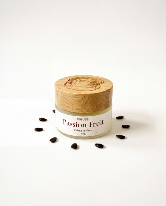 A small jar of WOUDH Passion Fruit Solid Crème Perfume with a light wood lid, set on a white background with scattered dark passion fruit seeds around it.