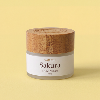 A 15g jar of WOUDH Sakura Crème Perfume with a light brown bamboo lid and a white label, set against a solid pale yellow background.